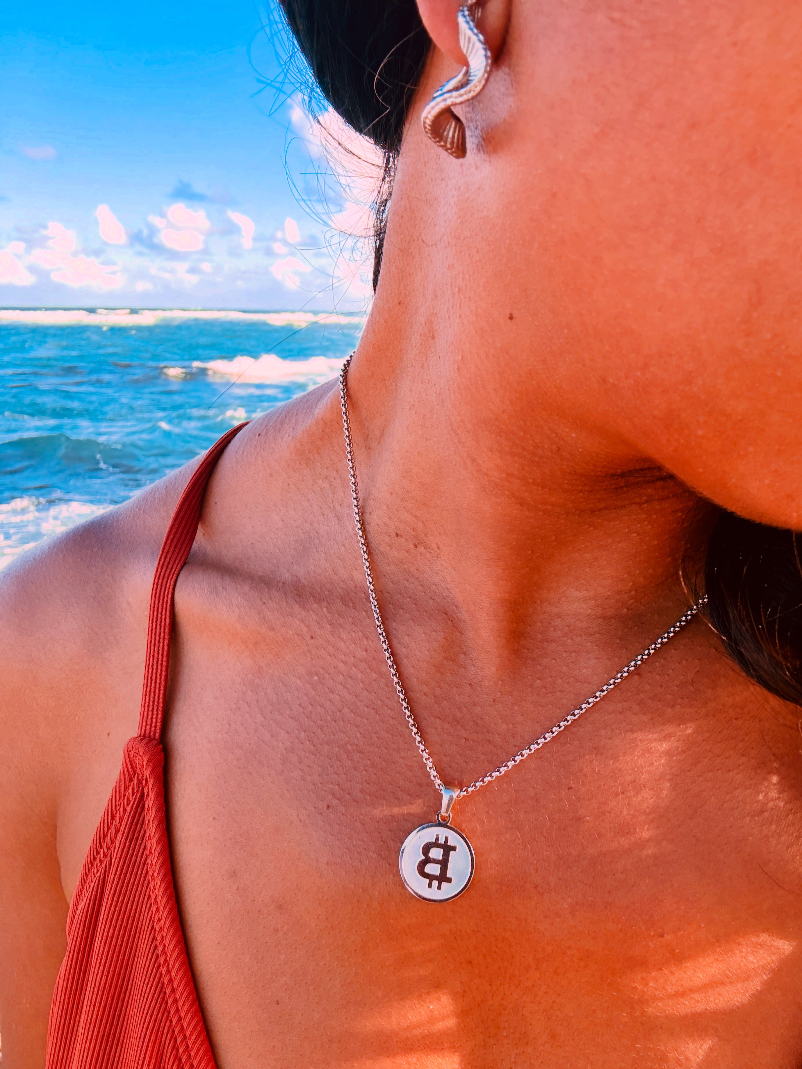 Person wearing a silver Bitcoin pendant necklace and hoop earrings with a beach in the background