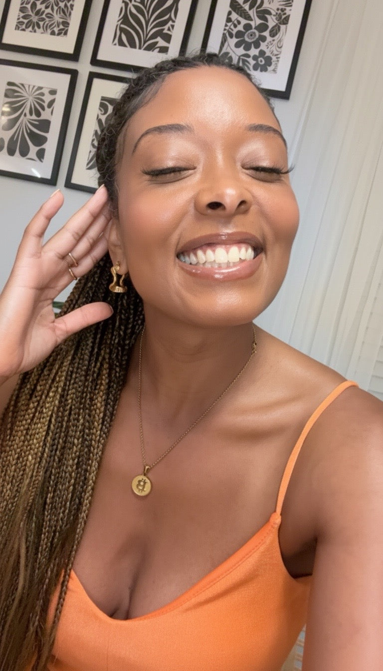 A smiling woman with long braided hair wearing a bright orange spaghetti-strap top, gold hoop earrings, and a gold pendant bitcoin necklace, and gold bit babe hoop earrings posing indoors against a wall with framed black-and-white floral art.